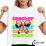 White T-shirt with a bright “Teacher” DTF transfer design featuring repeated colourful teacher text, a large pink bow, pencil, and eraser illustration.