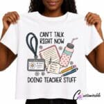 White T-shirt with “Can’t Talk Right Now Doing Teacher Stuff” DTF transfer design featuring teacher stationery, a notebook, pencils, ID badge, drink bottle, sticky note, and daisies.