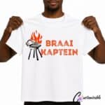 White T-shirt with “Braai Kaptein” DTF transfer design featuring red-orange text, a black braai grill illustration, and flame detail.