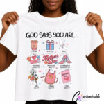 A mockup of a white custom t-shirt featuring a Christian Mother's Day design titled "GOD SAYS YOU ARE...". The layout displays a 3x3 grid of pastel-colored icons paired with encouraging traits and corresponding Bible verses, including a coffee cup for Strong (Isaiah 40:31), a gift box for Accepted (Romans 15:7), holding hands for Bold (1 John 5:14), a heart letter for Redeemed (Galatians 3:13), high heels for Courageous (Deuteronomy 31:6), a floral bouquet for Kind (Luke 6:27), an apron for Capable (Mark 10:27), and a card for Loyal (John 15:13), centered around a retro pink "Mother's Day" logo labeled Protected (2 Thess 3:3).