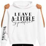 A mockup of a white pullover hoodie with a front pocket displaying a black DTF transfer design. The chest features the quote "LEAVE A LITTLE Sparkle" in a mix of elegant serif typography and cursive script, complete with small accent starburst sparkles. The right sleeve displays a vertical matching print that reads "WHEREVER you go" with small heart flourishes. The hoodie is held up by a model against a white background to clearly showcase the dual placement apparel print design.